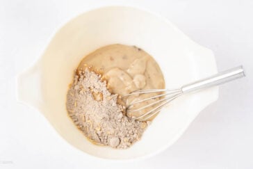 A white mixing bowl contains dry cake mix and condensed soup, partially blended with a metal whisk, on a white background.