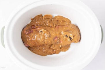 A white slow cooker containing two pieces of meat covered in a thick brown gravy with visible mushroom slices.