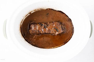 A cooked beef roast covered in brown gravy sits in a white slow cooker, surrounded by sauce with visible herbs and mushrooms.
