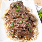 A rectangular white plate with shredded beef roast covered in brown mushroom gravy, garnished with parsley, served over a bed of brown rice.