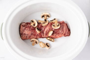 Raw beef and sliced mushrooms placed inside a white oval slow cooker, viewed from above against a light background.
