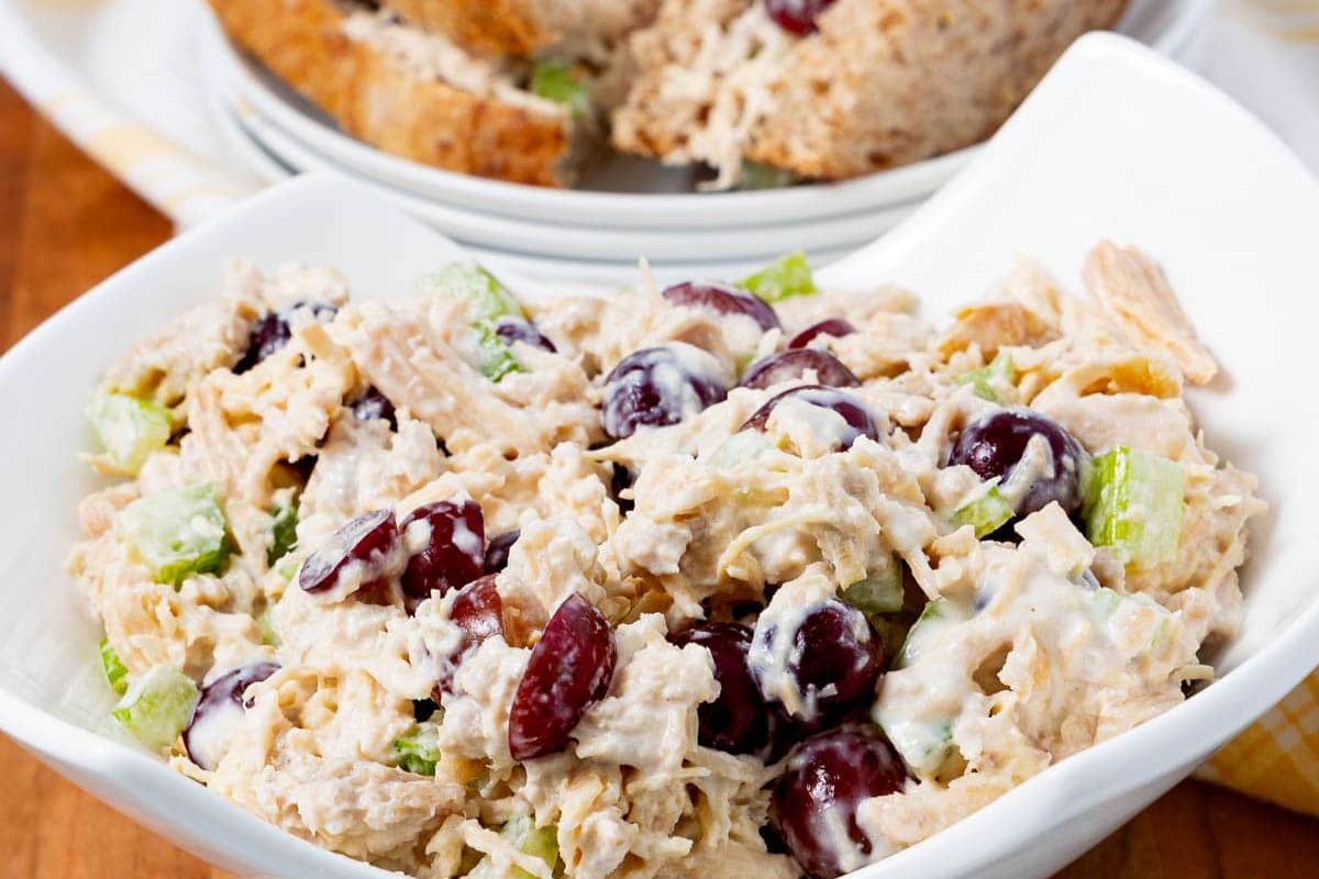 A close-up of chicken salad in a white bowl, featuring shredded chicken, halved red grapes, chopped celery, and creamy dressing. Slices of whole grain bread are visible in the background.