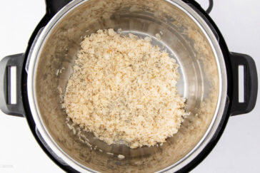 Uncooked white rice spread at the bottom of a stainless steel inner pot of an electric pressure cooker, viewed from above.
