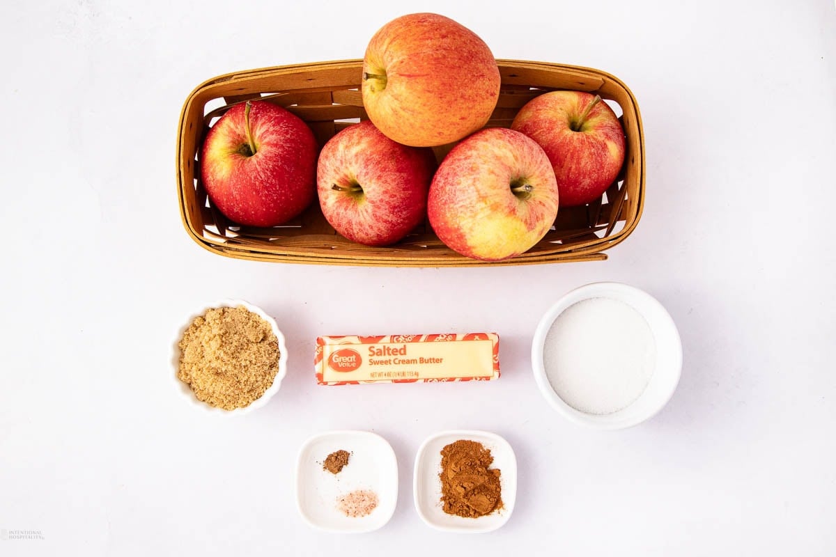 A basket with four apples, a stick of salted butter, a bowl of granulated sugar, a bowl of brown sugar, and two small dishes containing spices, all arranged on a white surface.