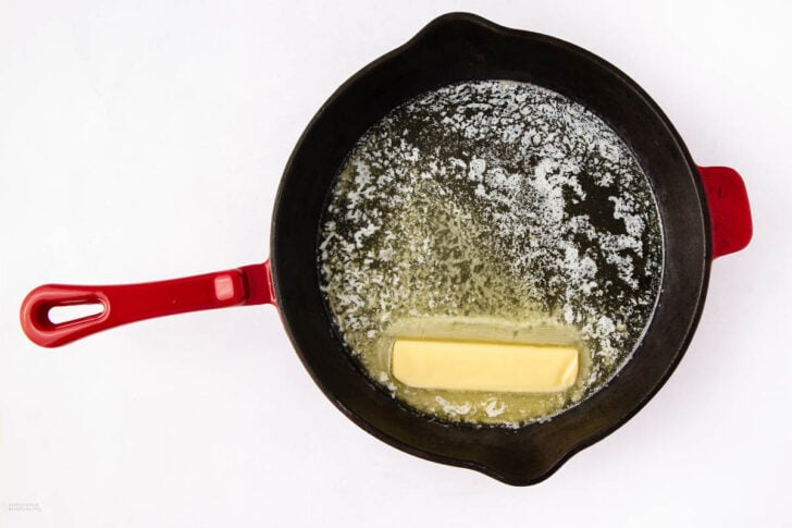A stick of butter melting in a black cast iron skillet with a red handle, photographed from above on a white background.