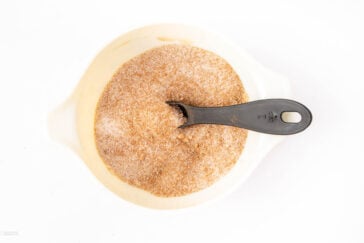A white bowl filled with a cinnamon and sugar mixture, with a black plastic tablespoon resting inside the bowl.