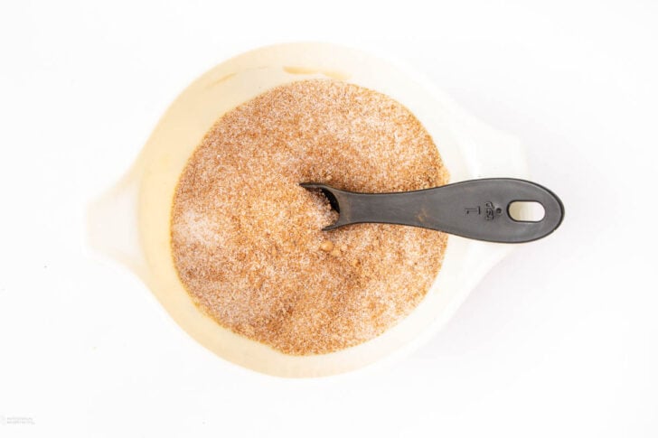 A white bowl filled with a cinnamon and sugar mixture, with a black plastic tablespoon resting inside the bowl.