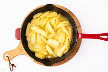 A black skillet with a red handle sits on a wooden board, filled with sliced apples cooking in a light syrup or sauce on a white background.