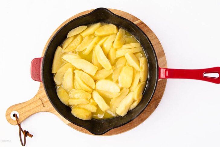 A black skillet with a red handle sits on a wooden board, filled with sliced apples cooking in a light syrup or sauce on a white background.