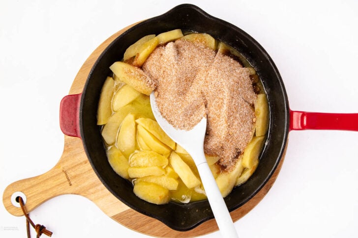 Sliced apples in a black skillet with melted butter, topped with a cinnamon-sugar mixture. A white spoon rests in the skillet, which sits on a wooden cutting board.