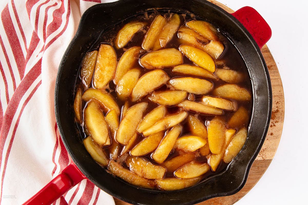 Sliced apples cooked in a skillet with a glossy brown cinnamon sauce, placed on a wooden board next to a red and white striped towel.