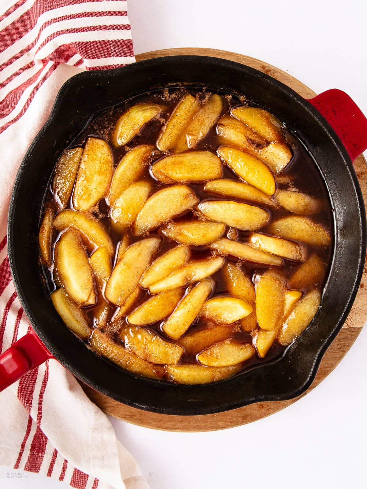 A black skillet filled with sliced apples cooked in a brown, syrupy sauce, placed on a wooden board next to a red-and-white striped kitchen towel.