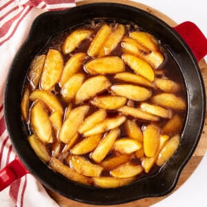 Sliced apples coated in a glossy brown cinnamon syrup, cooking in a black skillet with red handles, placed on a wooden board next to a white and red striped kitchen towel.
