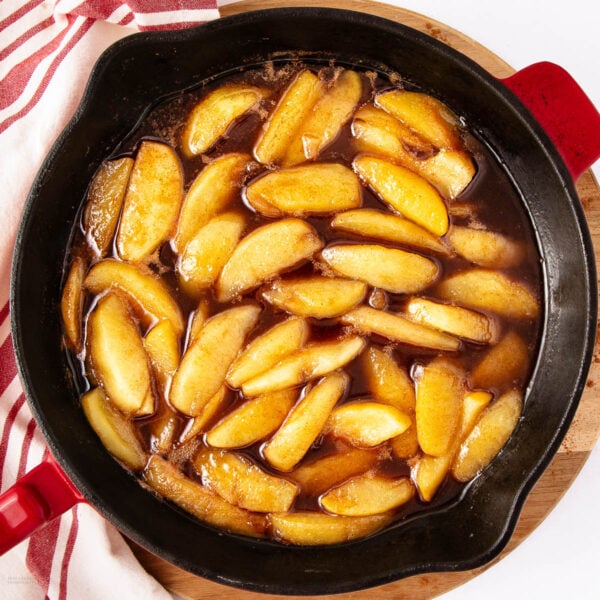 Sliced apples coated in a glossy brown cinnamon syrup, cooking in a black skillet with red handles, placed on a wooden board next to a white and red striped kitchen towel.