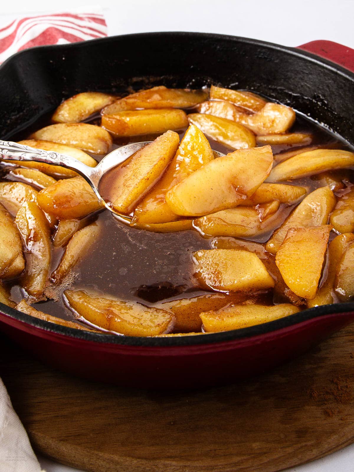 Sliced apples cooked in a cinnamon and brown sugar sauce are shown in a cast iron skillet with a spoon, resting on a wooden surface. A red and white cloth is partially visible in the background.