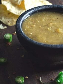 A small black bowl filled with green salsa sits on a dark surface. Sliced green chilies and broken tortilla chips are scattered around the bowl.