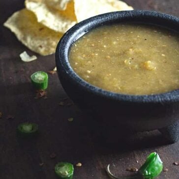 A small black bowl filled with green salsa sits on a dark surface. Sliced green chilies and broken tortilla chips are scattered around the bowl.
