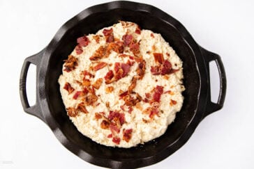A round cast iron skillet filled with a creamy white dip topped with crumbled pieces of bacon, set against a white background.