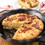 A cast iron skillet filled with golden brown cornbread topped with crispy bacon pieces, with one slice missing and some plates stacked in the background.