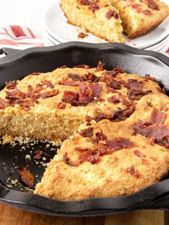 A cast iron skillet filled with golden brown cornbread topped with crispy bacon pieces, with one slice missing and some plates stacked in the background.