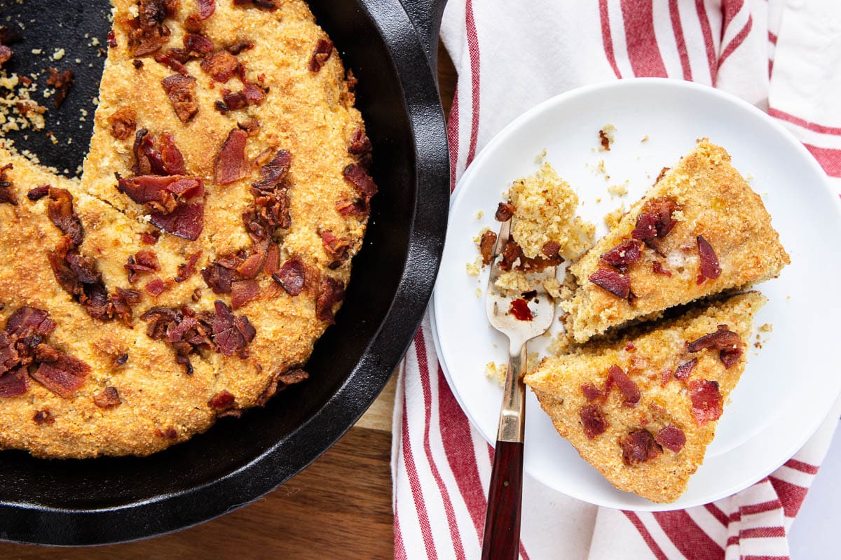 A skillet of cornbread topped with crispy bacon sits next to a white plate holding two slices of the cornbread, with a fork and a red-striped towel partially visible underneath.