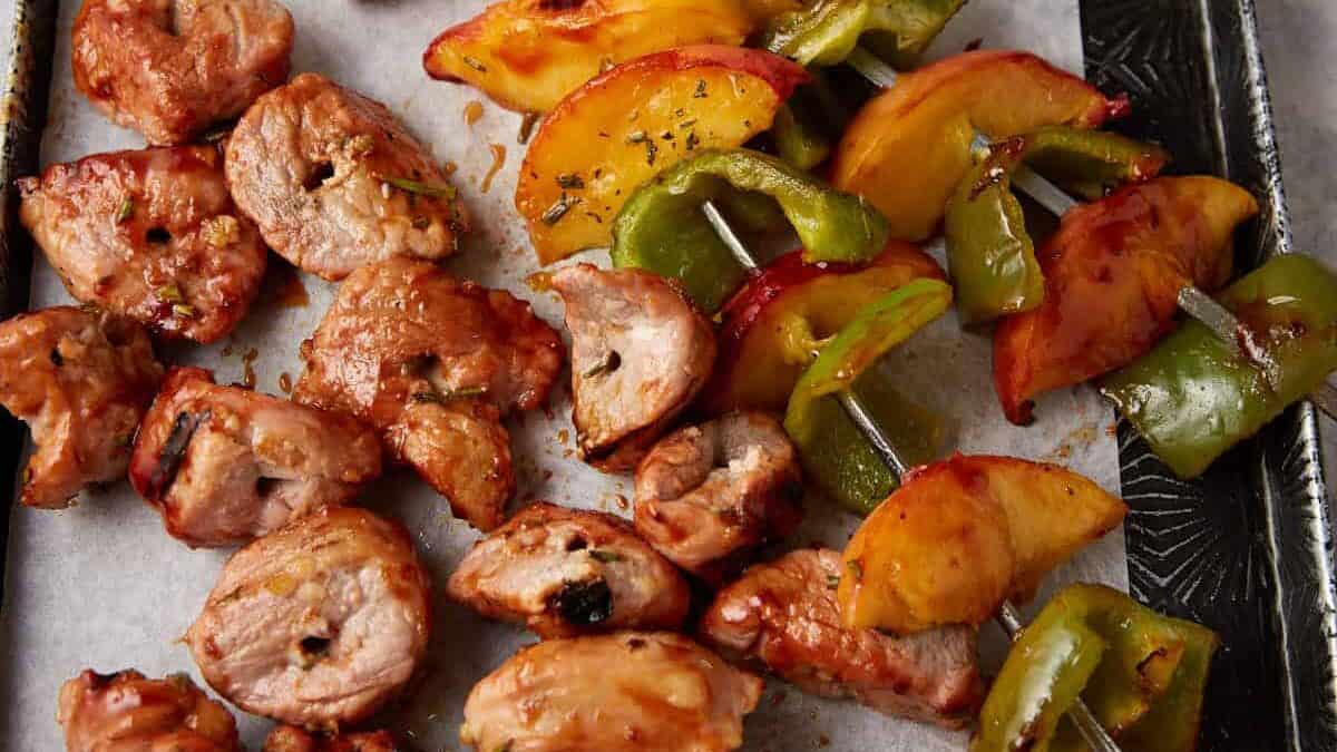 Grilled skewers of marinated meat, green bell peppers, and peach slices rest on a baking tray lined with parchment paper.