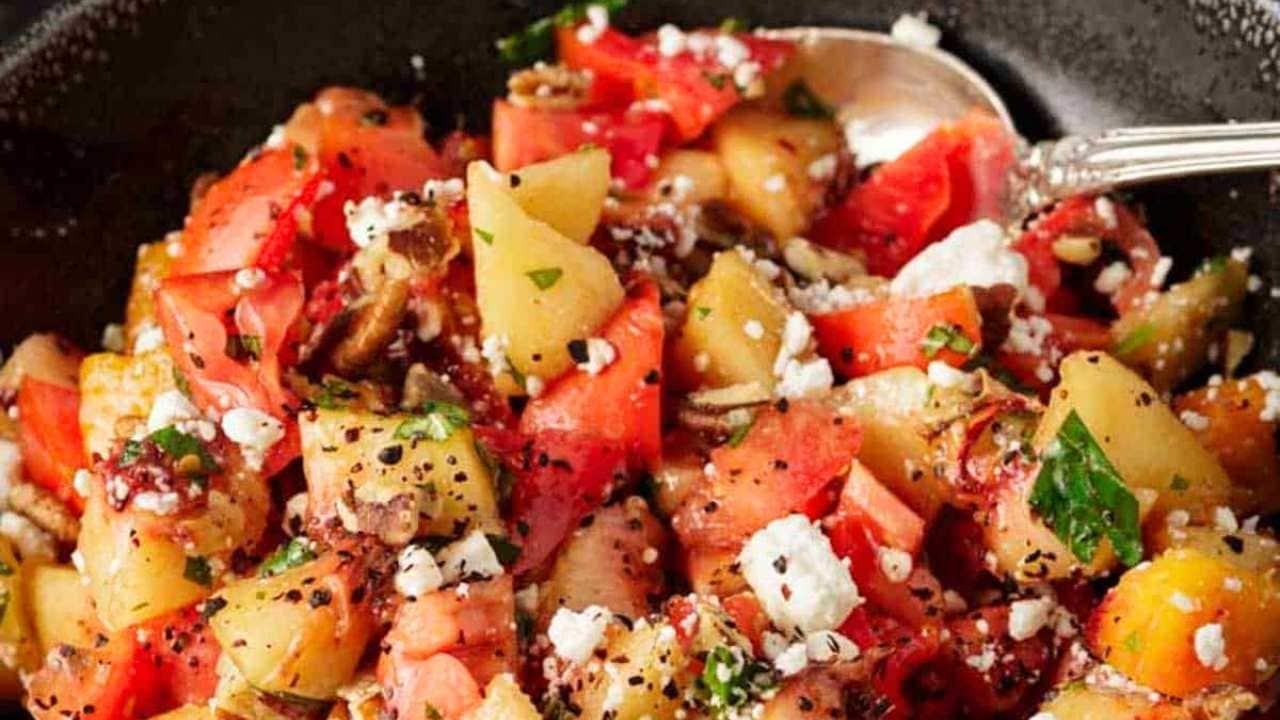 A close-up of a colorful salad with diced tomatoes and peaches, crumbled feta cheese, chopped herbs, and sprinkled black pepper, served in a dark bowl with a spoon.