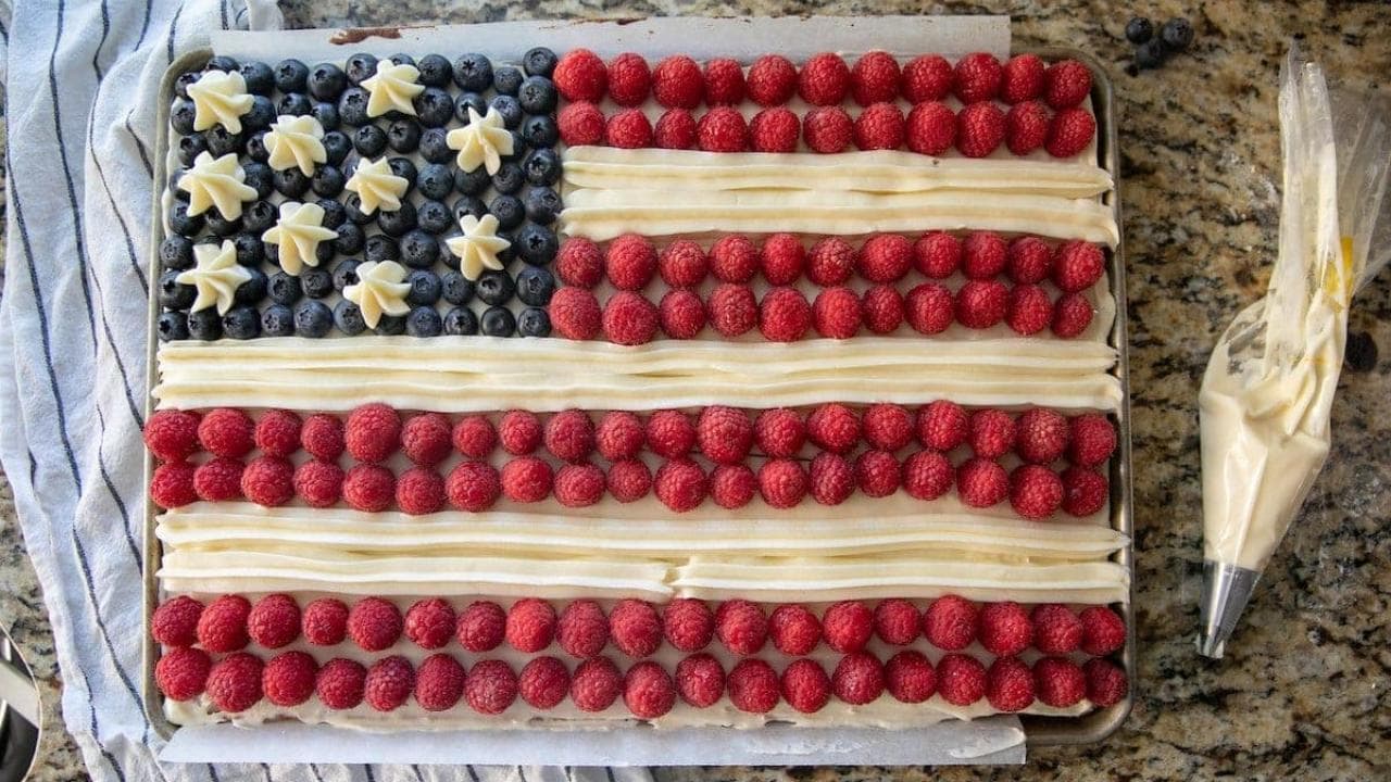 A rectangular cake decorated to look like the American flag, topped with blueberries, raspberries, white icing, and white chocolate stars, sits on a counter with a piping bag and towel nearby.