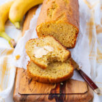 A loaf of banana bread on a wooden board, with two slices cut; one slice is spread with butter. Two bananas and a knife with a brown handle are beside the bread. White parchment paper lines the board.