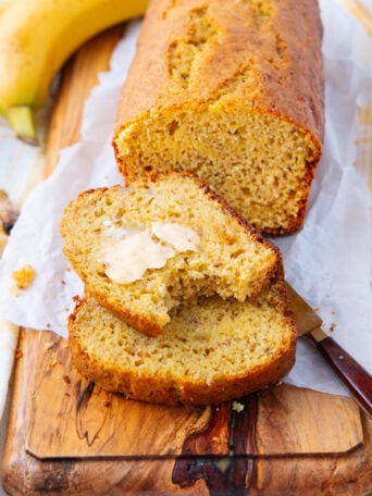 A loaf of banana bread on a wooden board, with two slices cut; one slice is spread with butter. Two bananas and a knife with a brown handle are beside the bread. White parchment paper lines the board.