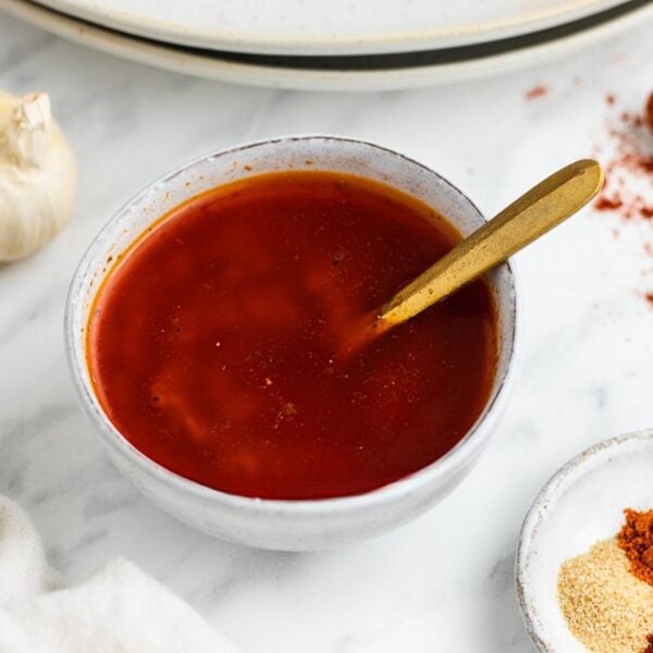 A small white bowl filled with red, oily enchilada sauce, with a gold spoon resting inside. Nearby, garlic and spices add flavor reminiscent of a canned cowboy candy recipe on a white marble surface.