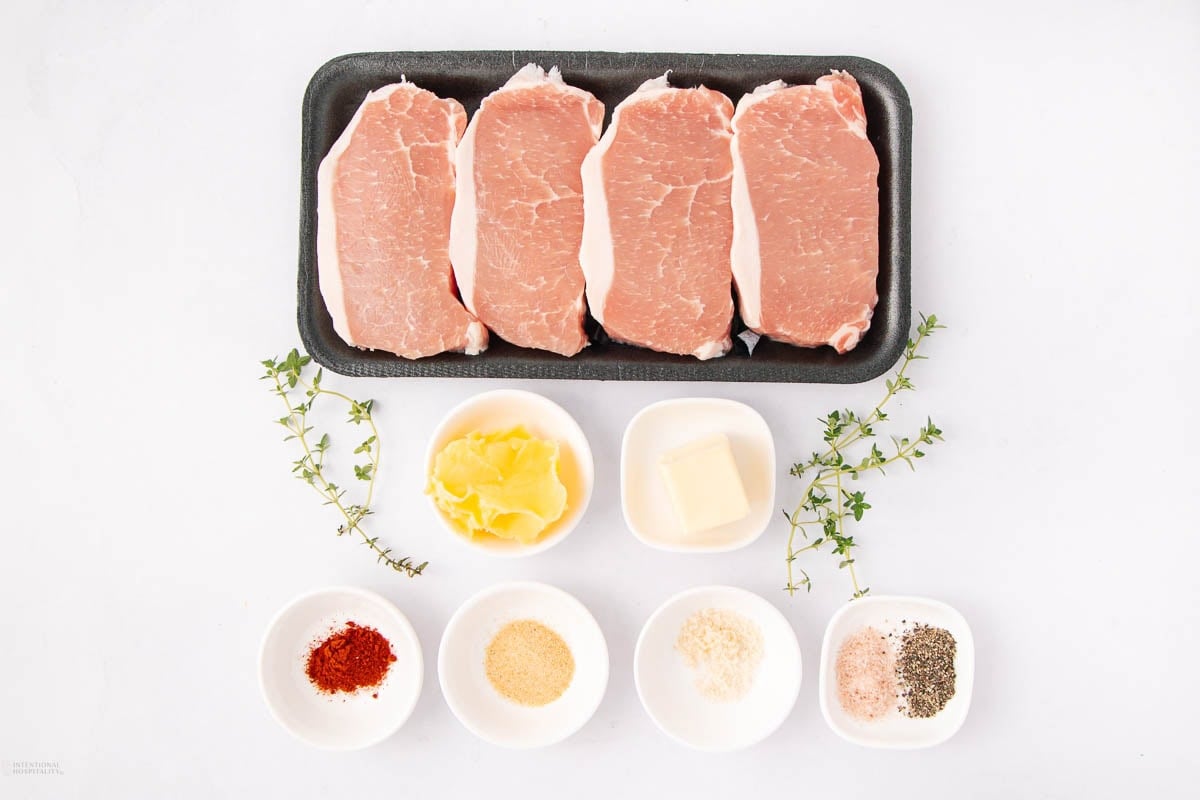 A tray with four raw pork chops is placed above small bowls containing spices, minced garlic, butter, grated Parmesan, salt, pepper, and paprika. Two sprigs of fresh thyme are also on the white surface.