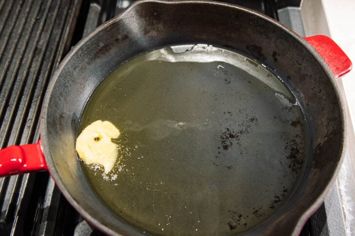A black cast iron skillet with a red handle sits on a stove, heating oil and melting a small piece of butter.
