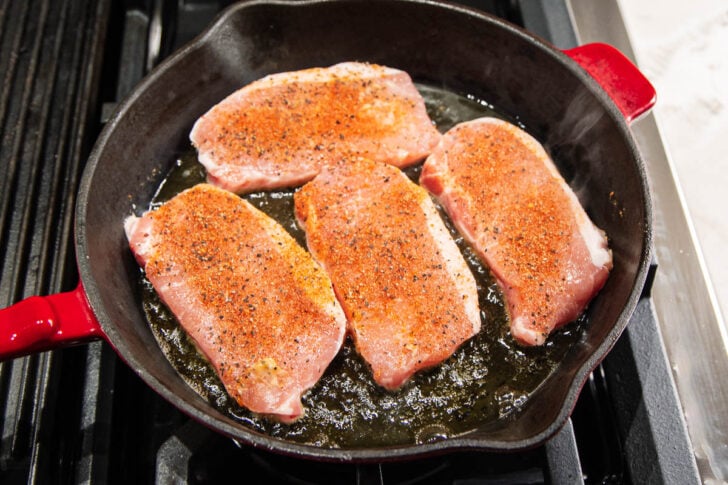 Four seasoned pork chops are sizzling in a black cast-iron skillet with a red handle, cooking on a stovetop. The meat is browning in hot oil, with visible spices and a slight steam rising.