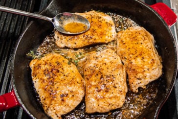 Four seasoned pork chops are searing in a cast-iron skillet with herbs and sizzling juices, while a spoon drizzles sauce over one of the chops.