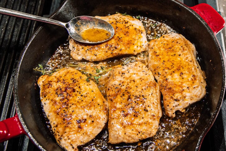 Four seasoned pork chops are searing in a cast-iron skillet with herbs and sizzling juices, while a spoon drizzles sauce over one of the chops.