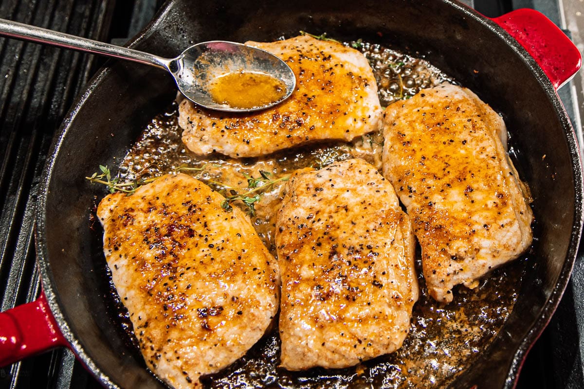 Four seasoned pork chops are searing in a cast-iron skillet with herbs and sizzling juices, while a spoon drizzles sauce over one of the chops.