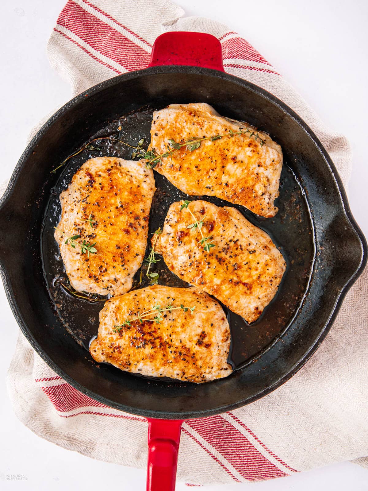 A cast iron skillet with four golden-brown seared pork chops garnished with fresh herbs, resting on a beige and red striped kitchen towel.