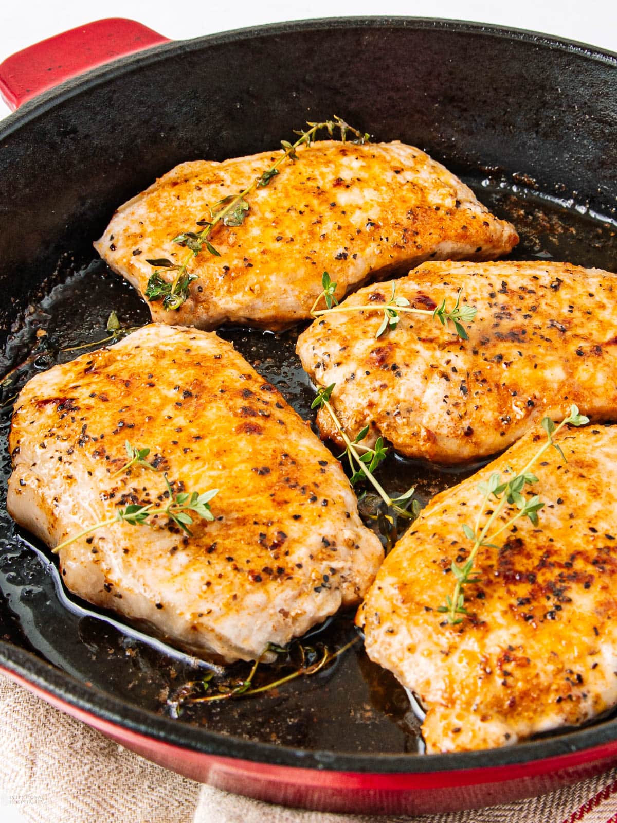 Four seasoned chicken breasts are being cooked in a black skillet, garnished with sprigs of fresh thyme. The chicken has a golden-brown sear, and the skillet sits on a beige cloth.