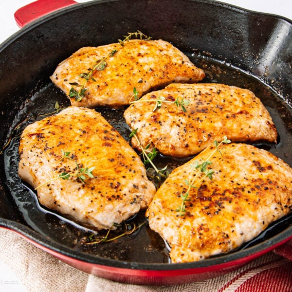 Four seasoned and seared chicken breasts cooking in a black cast iron skillet, garnished with fresh sprigs of thyme. The skillet rests on a red and white kitchen towel.