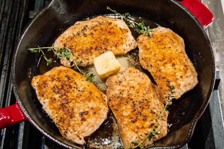 Four seasoned pork chops cooking in a cast iron skillet with a pat of butter melting in the center and sprigs of fresh thyme.