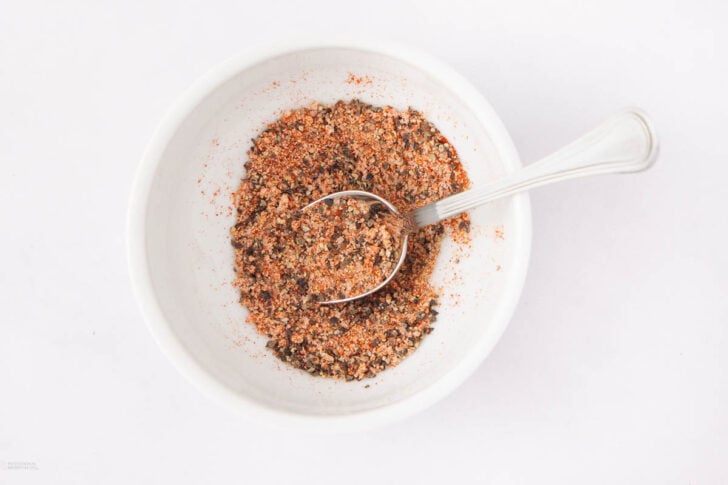 A white bowl filled with a coarse, reddish-brown seasoning mix, with a silver spoon resting inside the bowl. The bowl is placed on a plain white surface.