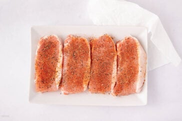 Four raw pork chops seasoned with spices and pepper are arranged in a row on a rectangular white plate. A folded white paper towel is placed in the upper right corner of the image.