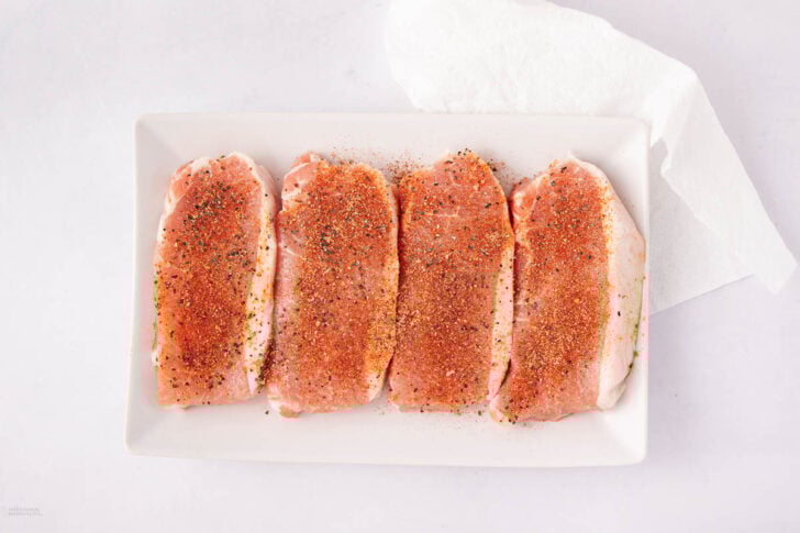 Four raw pork chops seasoned with spices and pepper are arranged in a row on a rectangular white plate. A folded white paper towel is placed in the upper right corner of the image.