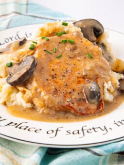 A plate of mashed potatoes topped with a pork chop, creamy mushroom gravy, and chives. The plate features a decorative inscription around the rim and rests on a blue plaid cloth.