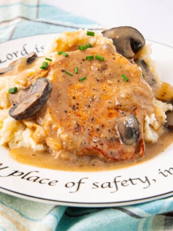 A plate of mashed potatoes topped with a pork chop, creamy mushroom gravy, and chives. The plate features a decorative inscription around the rim and rests on a blue plaid cloth.