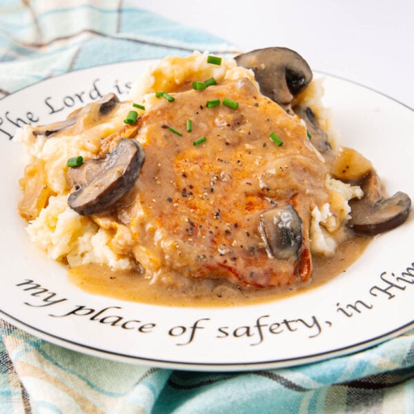 A plate of mashed potatoes topped with a pork chop, creamy mushroom gravy, and chives. The plate features a decorative inscription around the rim and rests on a blue plaid cloth.
