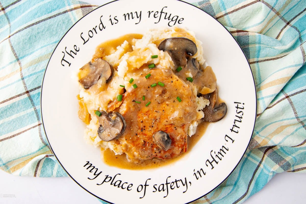 A plate with mashed potatoes topped with a pork chop, creamy mushroom gravy, and chives. The plate reads, "The Lord is my refuge, my place of safety, in Him I trust!" and rests on a blue plaid cloth.