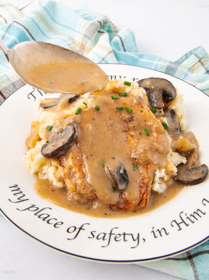 A plate of mashed potatoes topped with a Salisbury steak, brown gravy, sliced mushrooms, and chopped chives. A spoon drizzles extra gravy over the dish. A blue plaid napkin is in the background.