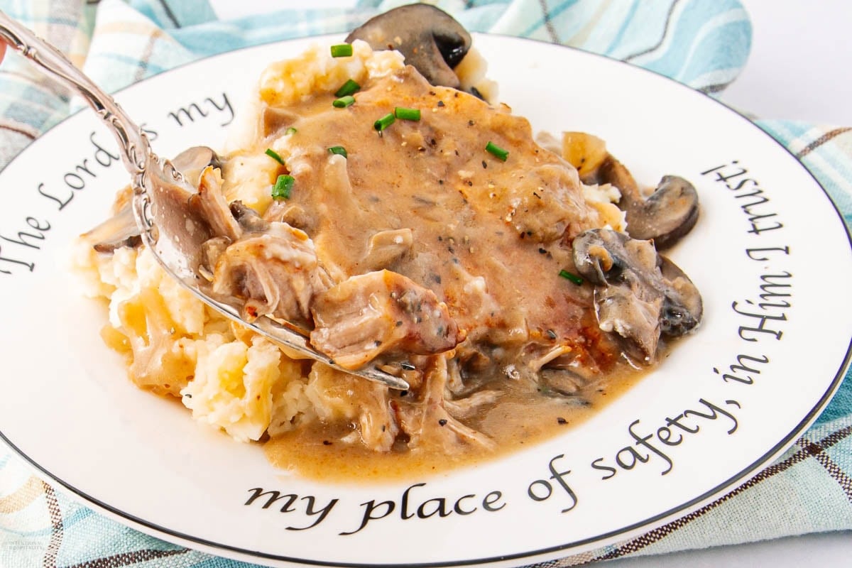 A plate of mashed potatoes topped with pork chops in creamy mushroom gravy, garnished with chopped chives. A fork rests on the plate, which has an inspirational quote on the rim. A blue and white cloth is underneath.